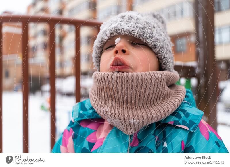 Kind in bunter Jacke fängt Schneeflocken mit offenem Mund vor einem Spielplatzhintergrund Offener Mund bunte Jacke Winter im Freien spielerisch kalt Wollmütze