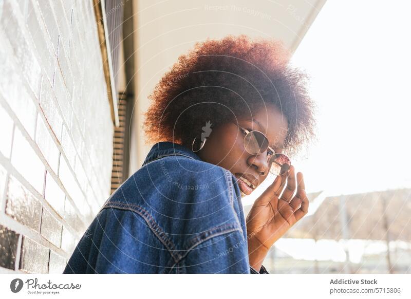 Eine nachdenkliche junge Frau rückt ihre runde Sonnenbrille zurecht, ihr Afro wird von der Sonne vor einer Backsteinmauerkulisse beschienen. beschaulich
