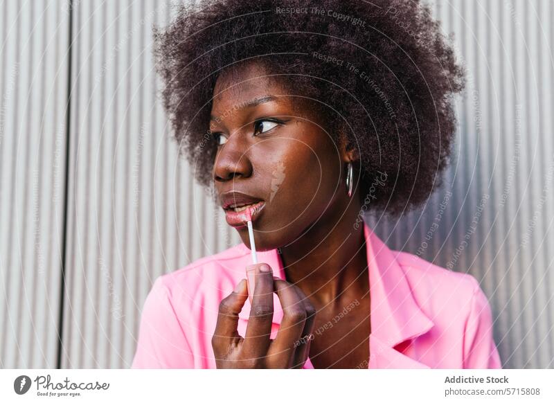 Eine modische schwarze Frau trägt mit Präzision Lipgloss auf, ihr Ausdruck ist konzentriert und unterstreicht einen Moment persönlichen Stils in einer städtischen Umgebung
