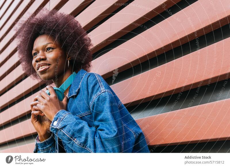 Eine strahlende junge Frau lächelt, Kopfhörer um den Hals, vor einem geometrischen Hintergrund, der urbane Coolness verkörpert Lächeln Jeansstoff Jacke cool