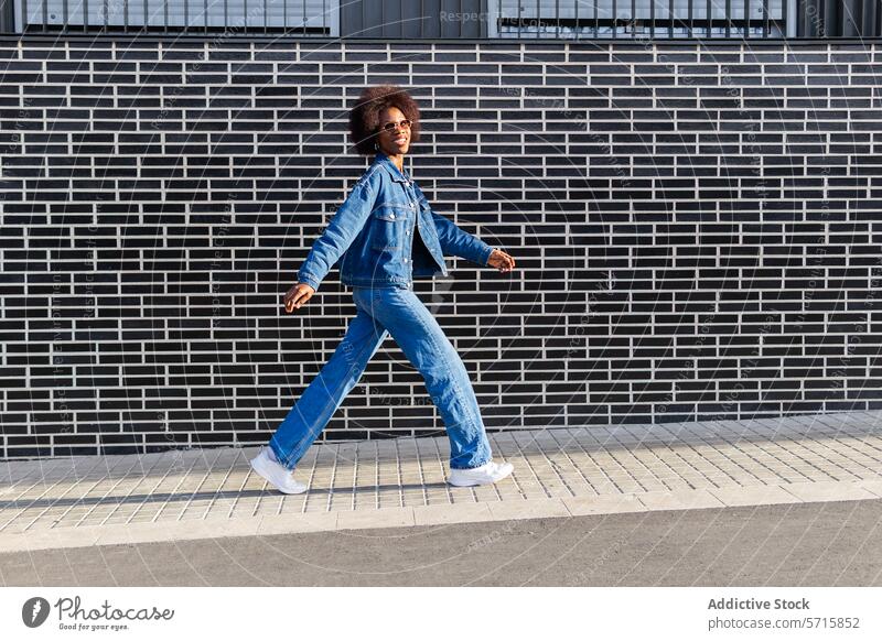Eine selbstbewusste afroamerikanische Frau in Jeans schreitet vorwärts, ihre dynamische Pose steht im Kontrast zu den grafischen Linien einer Backsteinmauer