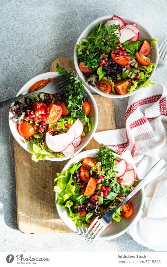 Draufsicht auf frische Gemüsesalate mit Kopfsalat, Rucola, Radieschen, Kirschtomaten und Granatapfelkernen auf einem Holzbrett mit einem Küchentuch Salatbeilage