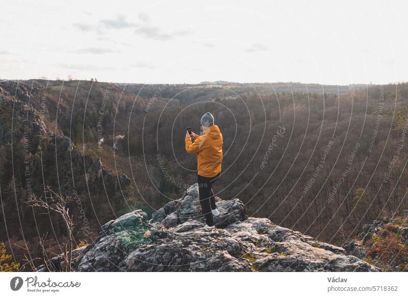 Ein Reisender in einer gelben Jacke, der am Rande einer Klippe steht und bei Sonnenuntergang mit seinem Mobiltelefon ein Foto von der Landschaft macht. Eine Erinnerung an eine Reise. Wildes Sarka-Tal, Prag