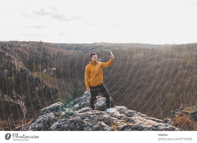 Ein Reisender in einer gelben Jacke steht am Rande einer Klippe und macht ein Selfie mit seinem Handy bei Sonnenuntergang.  Divoka Sarka Tal, Prag Lächeln
