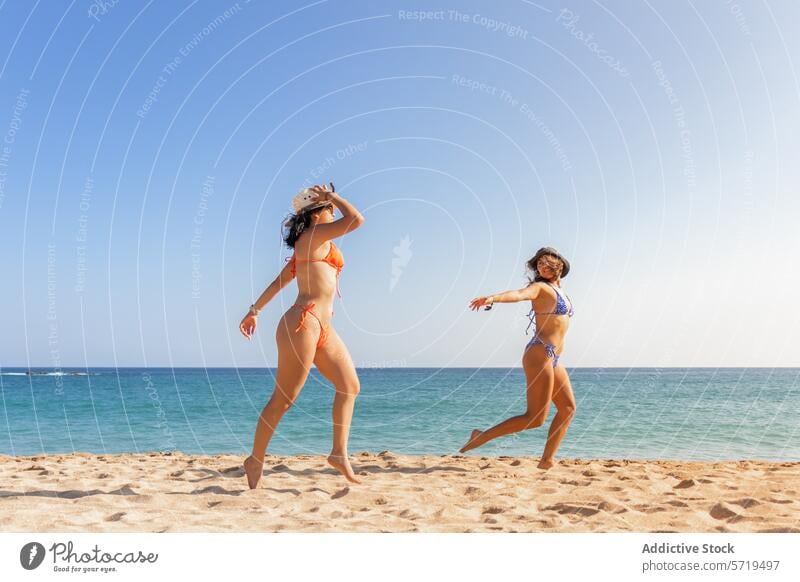 Zwei Freundinnen tanzen spielerisch auf dem Sand, tragen bunte Bikinis und Hüte und verkörpern so den Geist eines unbeschwerten Strandtages spielen Tanzen Hut