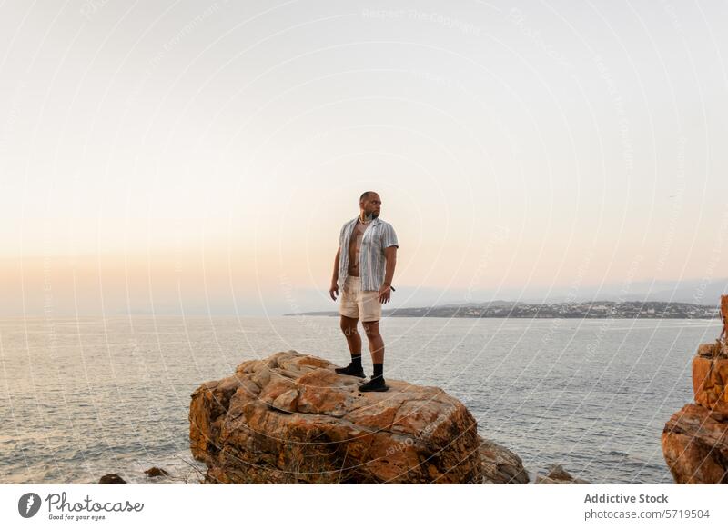 Ein Mann steht nachdenklich auf einer felsigen Klippe und blickt auf den weiten Ozean, während der Abendhimmel sanft in die Dämmerung übergeht Meer