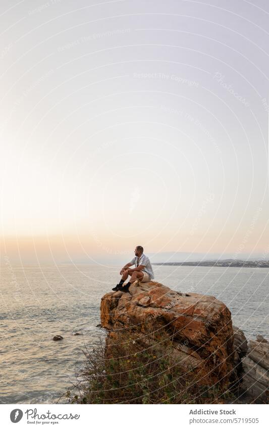 Eine Person sitzt auf einer zerklüfteten Klippe und blickt auf den Ozean hinaus, eingetaucht in die Ruhe eines sanften Sonnenuntergangs Einsamkeit