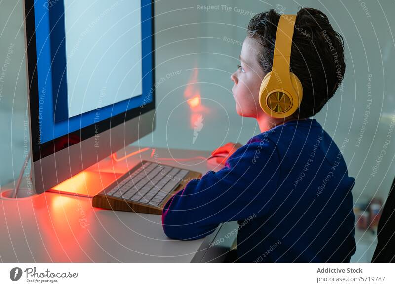 Ein Kind arbeitet konzentriert an einem Computer, beleuchtet von der warmen Hintergrundbeleuchtung des Monitors, mit Kopfhörern engagiert Technik & Technologie