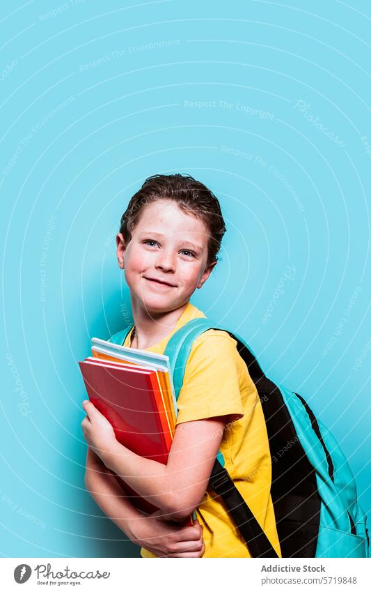 Ein Junge mit strahlenden Augen und einem aufrichtigen Lächeln trägt einen Stapel Bücher und seinen Rucksack und steht vor einem verspielten blauen Hintergrund