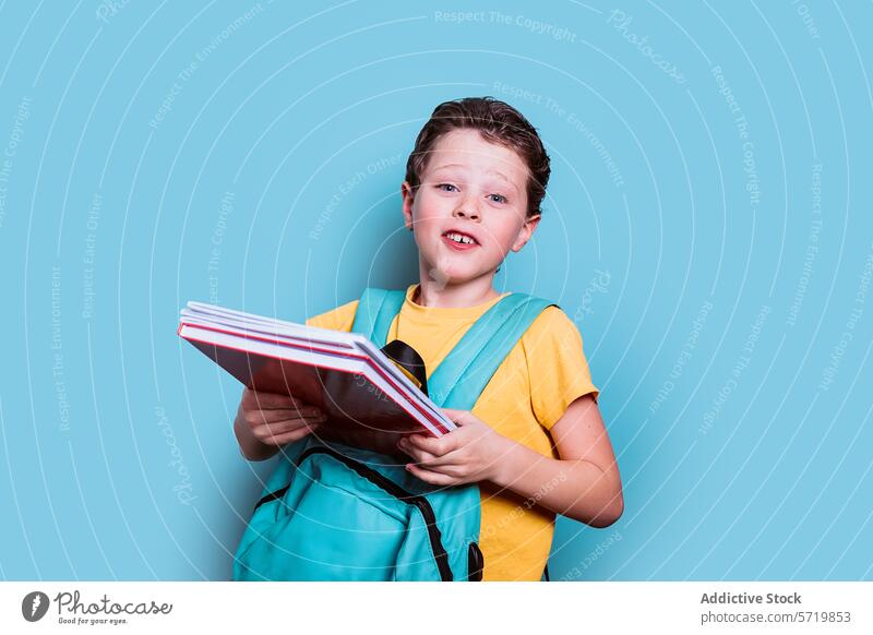 Ein Junge mit neugierigem Blick hält seine Schulbücher in der Hand, während er einen Rucksack trägt, vor einem erfrischend blauen Hintergrund Schule Bücher