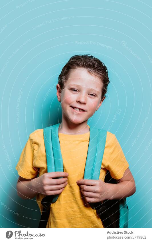 Ein fröhlicher Junge im Schulalter in einem gelben Hemd strahlt mit einem strahlenden Lächeln und hält sich an den Trägern seines Rucksacks fest, bereit für den Schultag
