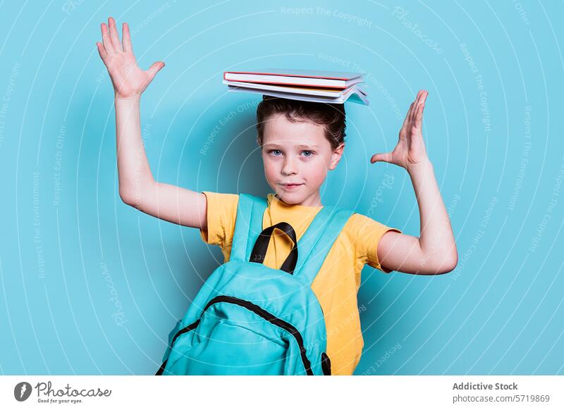 Ein Schuljunge mit Rucksack nimmt eine verspielte Pose ein und balanciert einen Stapel Bücher auf seinem Kopf vor einem hellblauen Hintergrund Junge Schule
