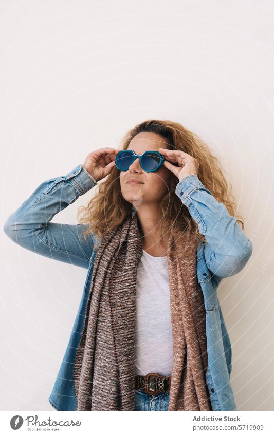 Eine stilvolle Frau in Jeansjacke und Strickschal rückt selbstbewusst ihre übergroße blaue Sonnenbrille vor einem schlichten Hintergrund zurecht Stil Jeansstoff