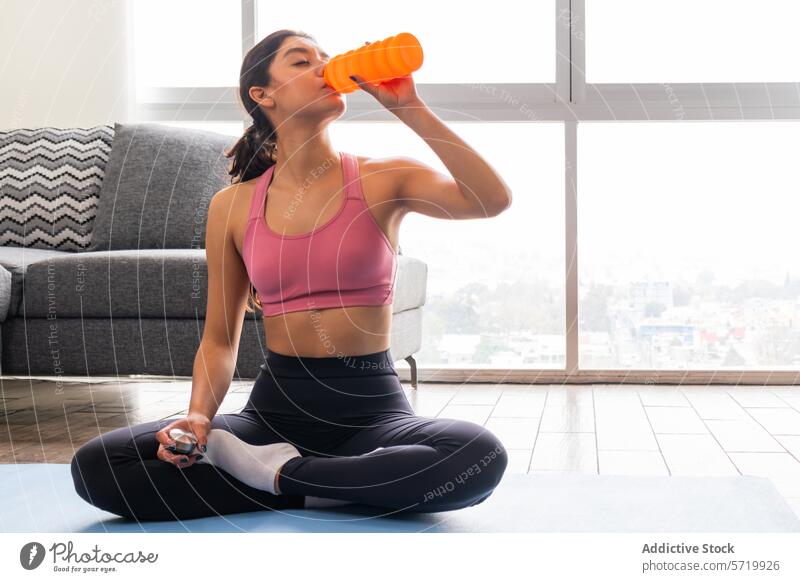 Trinkpause während der Yoga-Routine zu Hause Frau asiatisch Zwanziger Jahre Hydratation Wasserflasche orange sukhasana leichte Pose sitzend Yogamatte blau