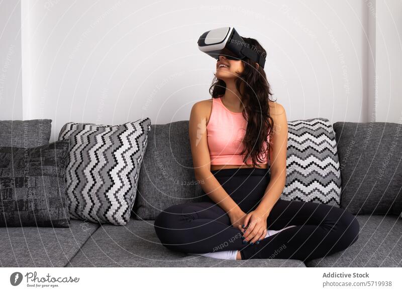 Frau mit VR-Headset auf Couch sitzend Virtuelle Realität Liege im Schneidersitz Fitness Yoga Meditation sportliche Kleidung freudig Wohnzimmer