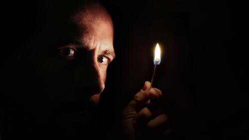 Besorgter Mann, der in der Dunkelheit mit einem Streichholz in die Kamera schaut. Konzept der Verdunkelung. Gesicht Filmriss Elektrizität Flamme Hand Licht