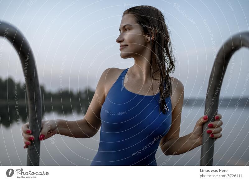 Ein atemberaubendes brünettes Mädchen in einem blauen Badeanzug nimmt an einem Sporttag teil. Trotz des kalten Wetters zeigt die schöne Schwimmerin ihre eleganten Kurven. Ihr strahlendes Profil verleitet dazu, in das tiefe, dunkle Wasser einzutauchen.
