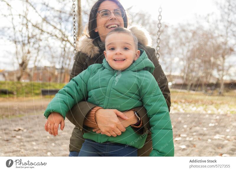 Ein kleiner Junge strahlt vor Glück auf einer Schaukel, sicher gehalten von seiner Mutter, beide genießen einen schönen Tag im Park pendeln Fröhlichkeit