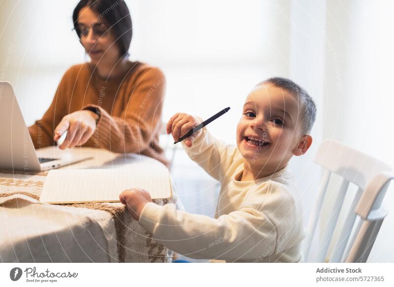 Ein fröhlicher Junge grinst in die Kamera und hält einen Stift in der Hand, während er mit seiner Mutter an einem Tisch sitzt, die auf ihren Laptop konzentriert ist