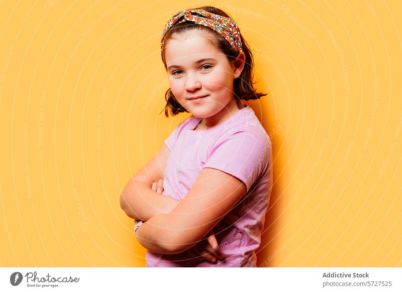 Selbstbewusstes junges Mädchen, das mit verschränkten Armen vor einem gelben Hintergrund posiert Kind selbstbewusst Pose die Arme verschränkt T-Shirt Stirnband