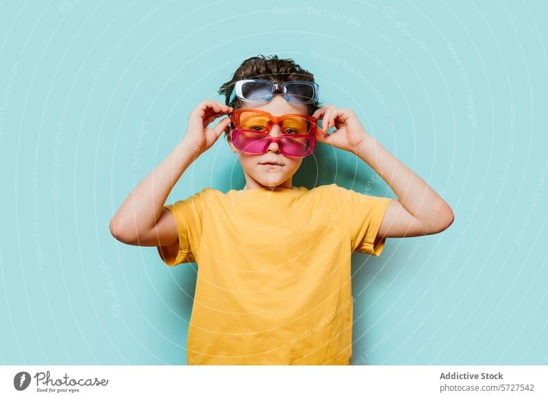 Kind mit mehreren Sonnenbrillen auf blauem Hintergrund Junge gelb Hemd farbenfroh Mode Stil schrullig Accessoire spielerisch Spaß Brille Stehen Kreativität