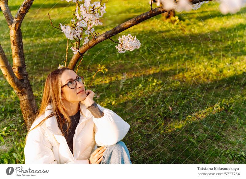 Schöne junge Frau in weißer Jacke und Brille sitzt in der Nähe blühenden Baum Garten Natur Kirsche im Freien Blüte Überstrahlung Schönheit Park schön lang