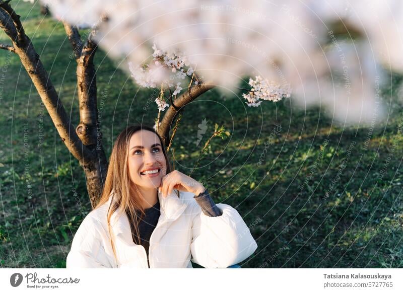 Schöne Frau in einer weißen Jacke sitzt unter blühenden Baum im Stadtpark im Frühling jung Garten Natur Kirsche im Freien Blüte Überstrahlung Schönheit Park