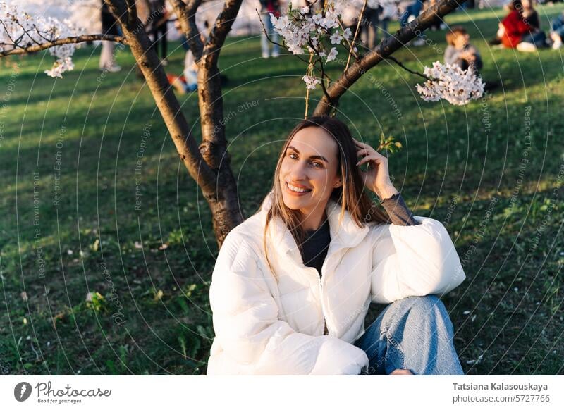 Lächelnde fröhliche Frau in weißer Daunenjacke und Jeans sitzt auf Gras im Stadtpark im Frühling unter Kirschblüte jung Garten Natur Kirsche im Freien Blüte
