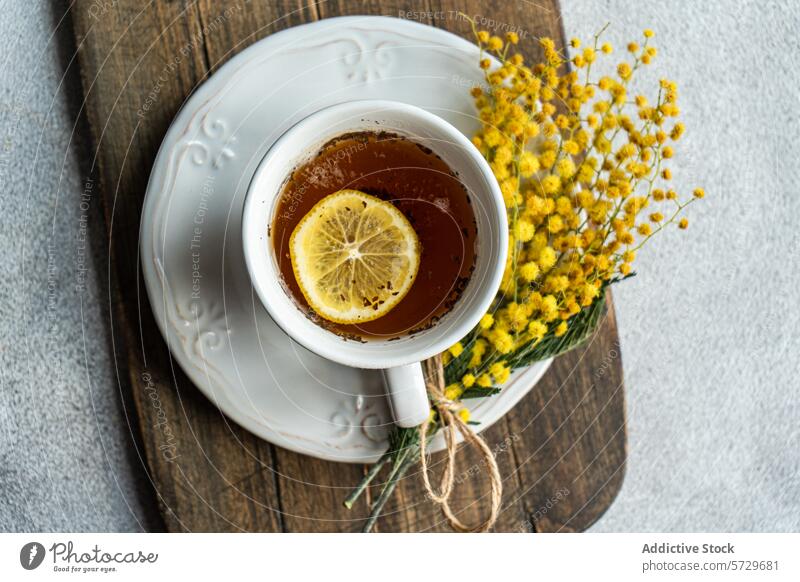 Draufsicht auf eine einladende Tasse Tee mit einer leuchtenden Zitronenscheibe auf einer verzierten Untertasse, begleitet von einem zarten Mimosenblütenbündel, präsentiert auf einem Holzbrett mit einem strukturierten Hintergrund