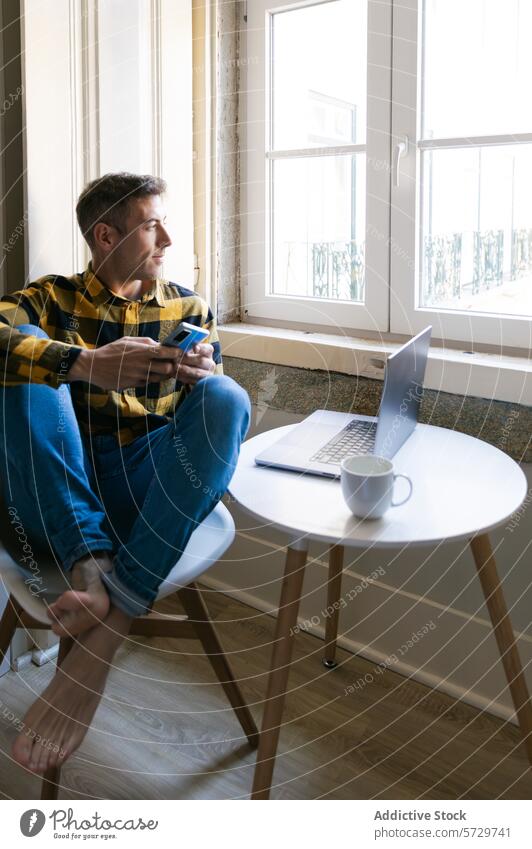 Mann genießt die Kaffeepause bei der Arbeit zu Hause Pause arbeiten heimwärts Barfuß Fenster schlürfend Smartphone Laptop Tisch im Innenbereich Erholung