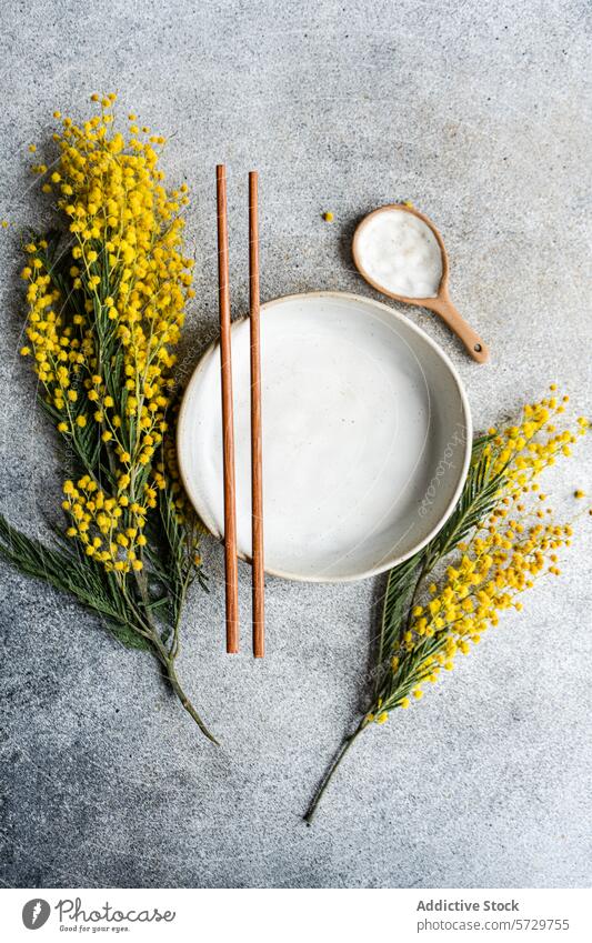 Draufsicht auf einen eleganten, minimalistisch gedeckten Tisch mit einem Keramikteller und Essstäbchen, akzentuiert durch das leuchtende Gelb der Mimosenblüten vor einem strukturierten grauen Hintergrund