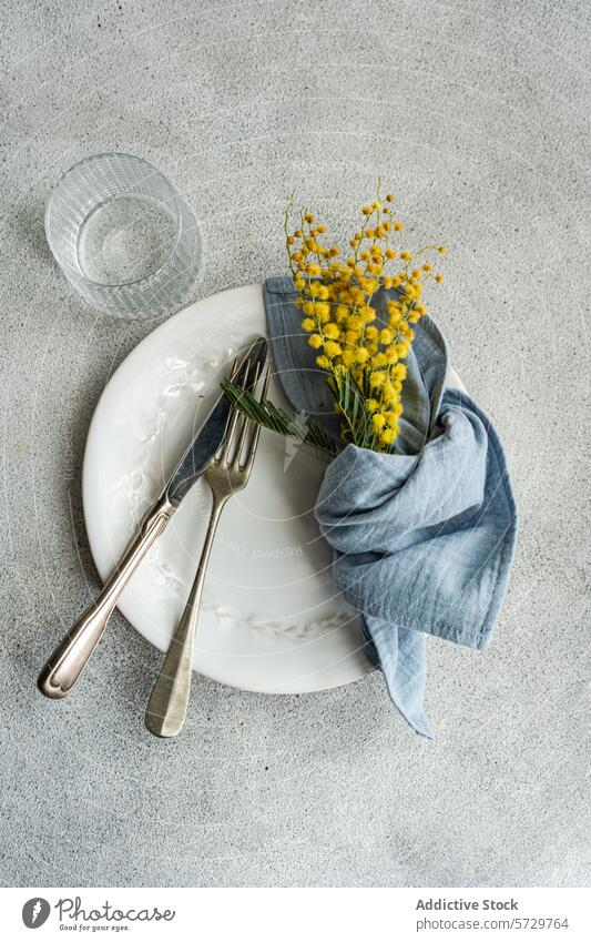 Elegante Tischdekoration mit einem weißen, verzierten Teller, altem Silberbesteck, einer blauen Leinenserviette und einem leuchtend gelben Mimosenstrauß auf einem strukturierten Hintergrund