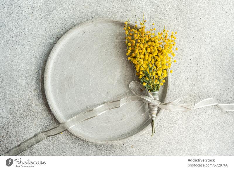 Draufsicht auf ein zartes Arrangement aus leuchtenden Mimosenblüten, gebunden mit einem silbernen Band, präsentiert auf einer glatten, strukturierten Tischoberfläche für einen raffinierten Touch