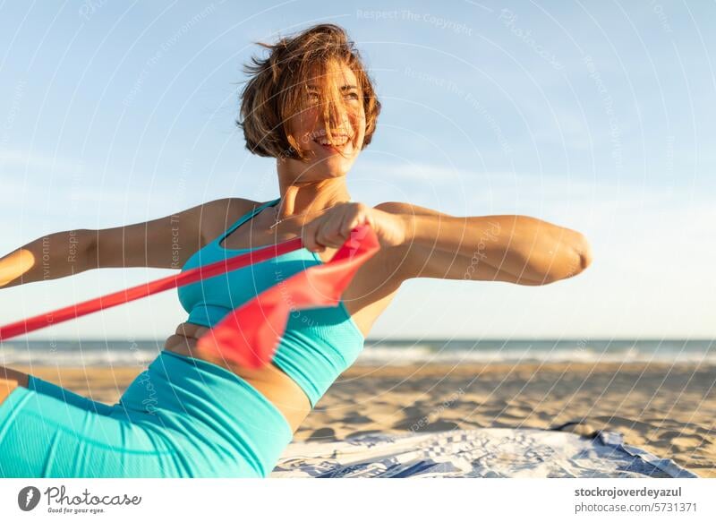 Eine Frau lächelt glücklich, während sie am Strand, in der Sonne und im Freien, mit einem Gummiband Pilates-Übungen macht Yoga mediterran Spanien