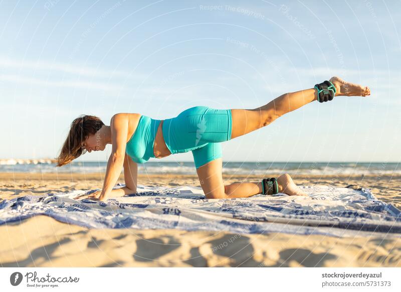 Eine Frau führt beim Pilates am Strand eine Beinzugbewegung mit zusätzlichem Gewicht in den Knöcheln aus Übung Yoga mediterran Spanien Körper-Geist-Übung