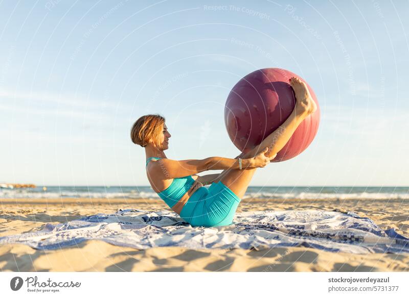 Eine Frau übt am Strand mit einem Pilates-Ball die Bewegung der doppelten Beinstreckung abdominal Beine Übung Yoga mediterran Spanien Körper-Geist-Übung