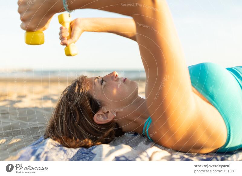 Eine Frau führt am Strand liegend Dehnungs- und Entspannungsübungen mit leichten 1 kg Hantelgewichten durch. Kurzhantel Pilates Yoga mediterran Spanien