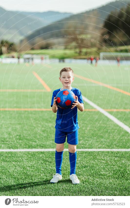 Junger Fußballspieler hält einen Ball auf dem Feld Spieler Uniform Sport Platz Jugend Kind sportlich aktiv im Freien Beteiligung Lächeln Selbstvertrauen Gras
