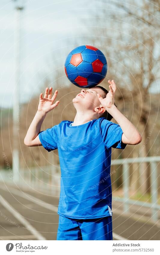 Junger Junge balanciert Fußball auf dem Kopf im Freien Ball Ausgewogenheit Sport Uniform blau rot entschlossen Ausdruck übersichtlich Himmel Hintergrund jung