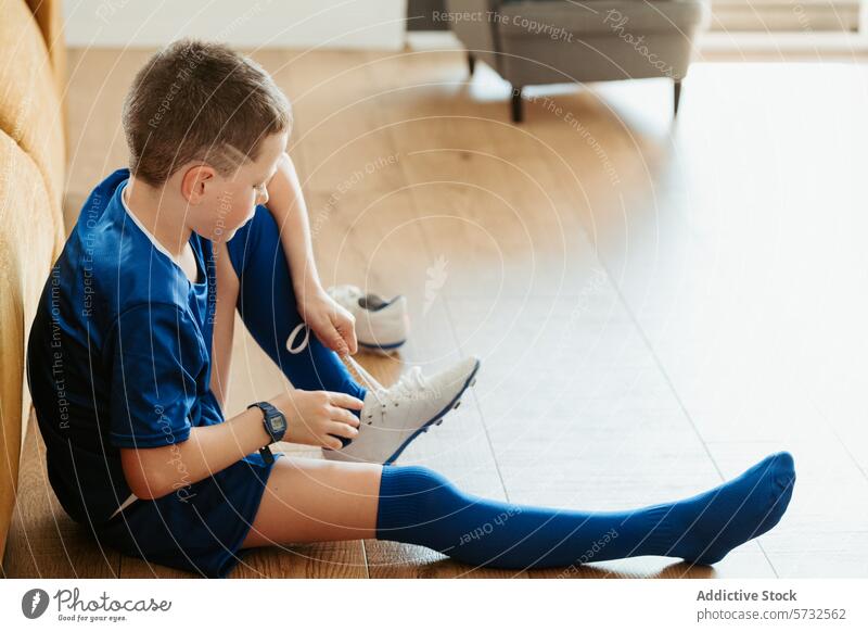 Junge in Fußballkleidung bindet Schnürsenkel im Haus Uniform Sport Schuh Kopplungsbindung Sitzen Stock hölzern im Innenbereich blau weiß Kind Sportbekleidung