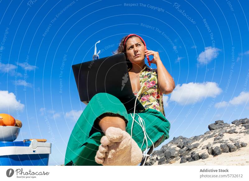 Von unten betrachtet führt eine fokussierte digitale Nomadin ein Geschäftsgespräch mit einem Laptop auf den Knien, vor einem Hintergrund aus schroffen Strandfelsen und blauem Himmel