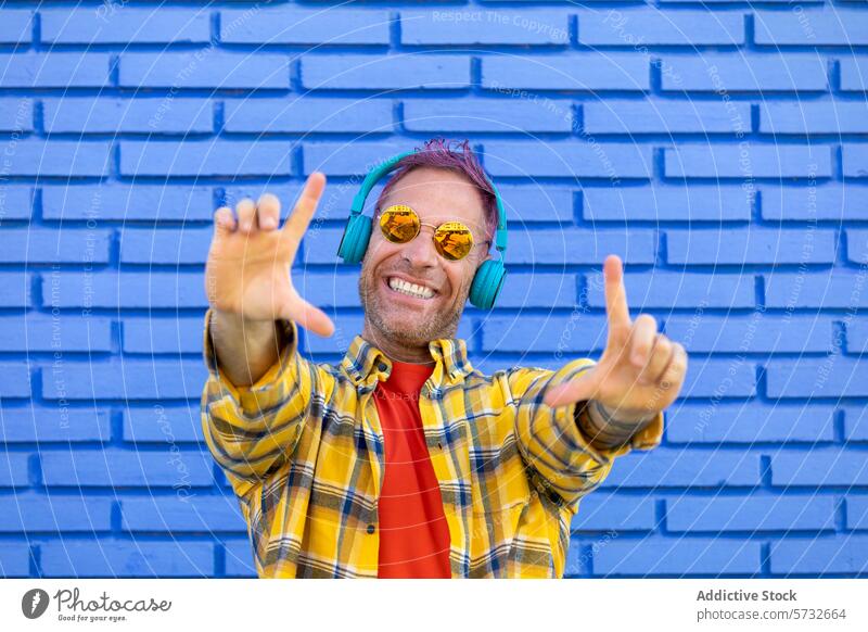 Ein verspielter Mann mit bunten Kopfhörern wirft ein Rock-Schild hoch und genießt die Musik mit einer blauen Backsteinmauer als lebendige Bühne Riffel schlagen