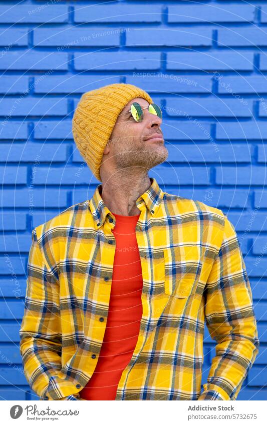 Ein fröhlicher Mann mit gelber Mütze und Sonnenbrille blickt auf und sonnt sich im Sonnenlicht vor einer strukturierten blauen Backsteinwand im Hintergrund.