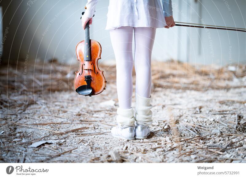 Kleines Mädchen ganz in Weiß wie eine Ballerina gekleidet, steht inmitten einer Baustelle mit dem Rücken zum Fotografen und hält eine hölzerne Violine Geige in der Hand, um im kreativen Chaos zu musizieren Musik zu spielen