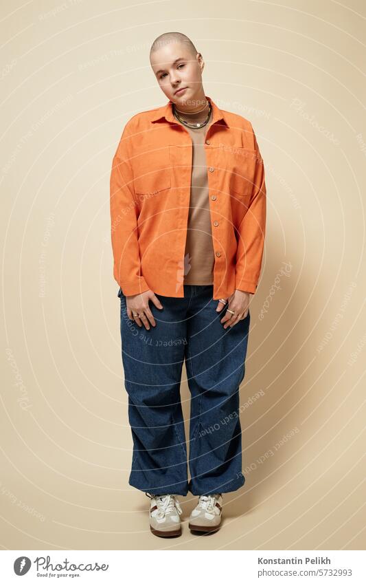 Vertikales Ganzkörperporträt einer jungen Frau mit Glatze, die eine leuchtend orangefarbene Jacke und weite Jeans trägt und in einem Studio posiert und in die Kamera schaut