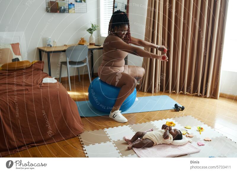 Glückliche Mutter eines neugeborenen Babys beim Training zu Hause Kurzhantel Übung heimwärts aktiv Lifestyle Fitnessball Sitz positionieren Mama Kind spielen