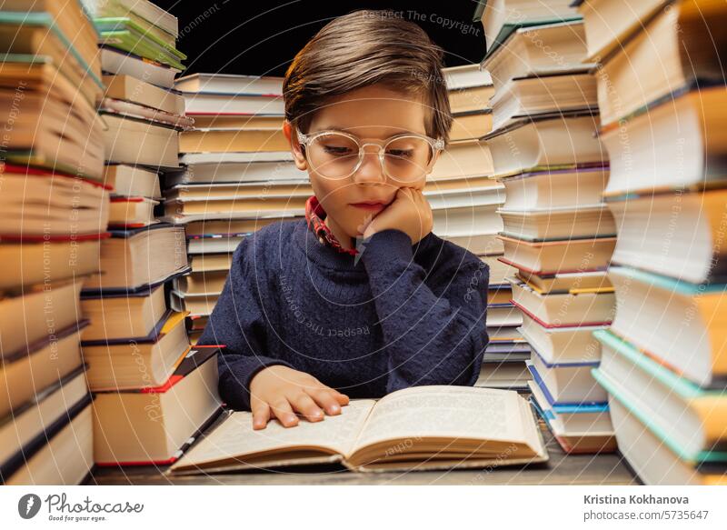 Niedliche kleine Schüler Junge in Brille Lesen interessantes Buch in der Bibliothek zwischen Stapeln von Büchern Literatur. Bildung Konzept, Vorbereitung oder Grundschule