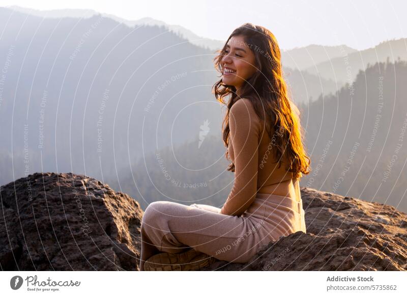 Eine fröhliche Frau sitzt im goldenen Licht des Sonnenaufgangs auf einem Berggipfel, mit einer atemberaubenden Kulisse von nebligen Bergen Freude Gelassenheit