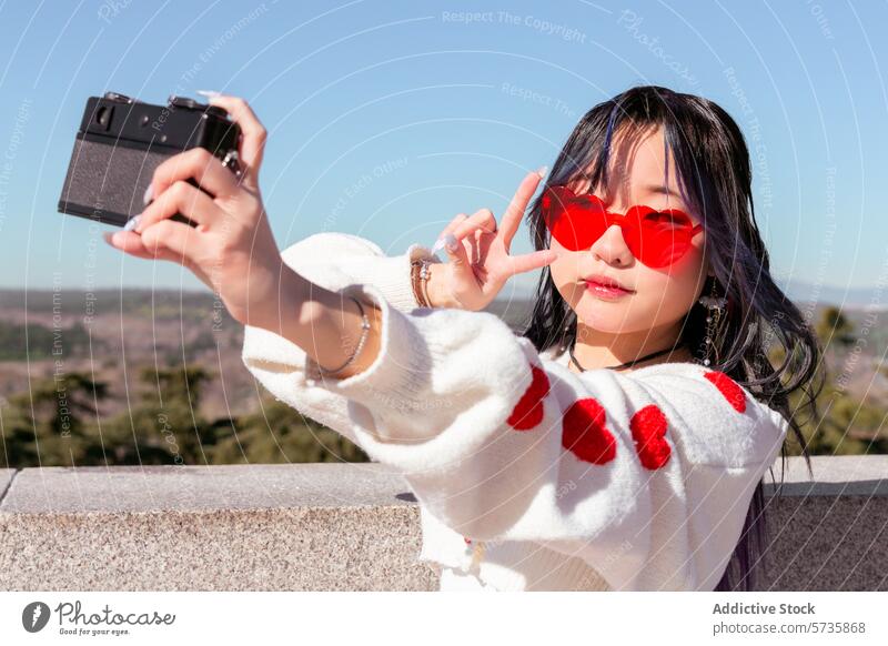 Junges chinesisches Mädchen nimmt Selfie mit Vintage-Kamera im Freien Chinesisch Oldtimer-Kamera Gen-Z herzförmige Sonnenbrille Pullover Herzen jung retro