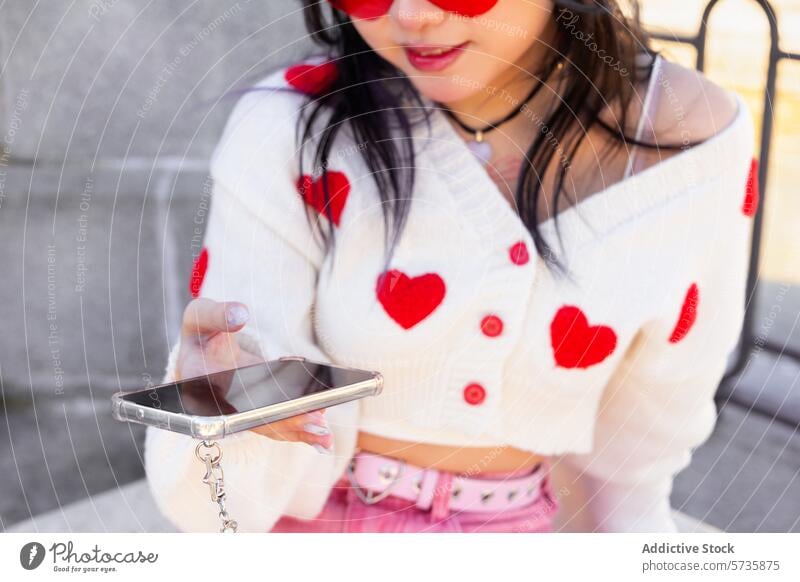 Modisches Gen-Z-Girl mit Smartphone und Herz-Strickjacke Mädchen Model Stil Jugend trendy Sonnenbrille modern lässig im Freien posierend Technik & Technologie
